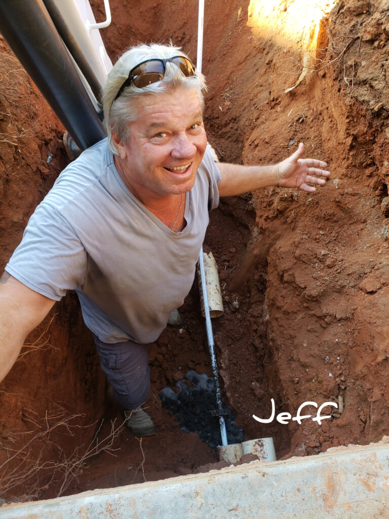 Man smiling in a narrow dirt trench holding a shovel